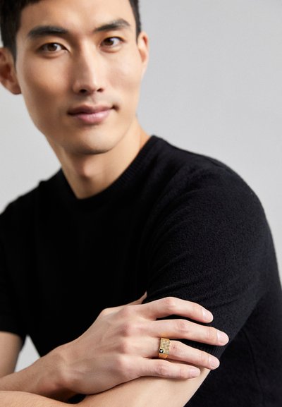 Gold ring with a grid pattern and dark accent detail, worn on a hand resting on an arm. Model wears a black sweater.