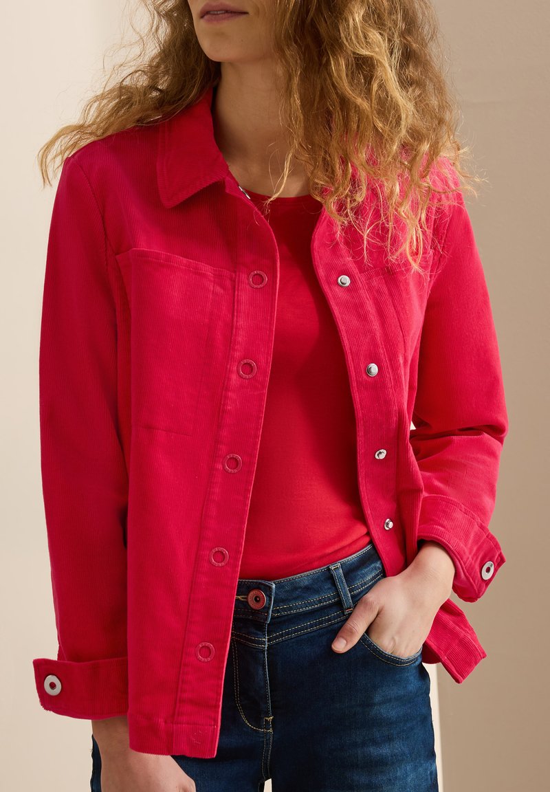 Röd corduroyjacket med metallknappar, en bröstficka och uppvikta ärmar, kombinerad med en röd topp och denimjeans.