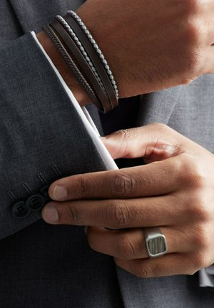 Leren armbanden in donkerbruin en grijstinten, gestapeld op een hand, met geweven en gladde texturen. Een zilveren ring met een houten accent.