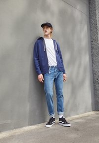 Granatowa bluza z zamkiem błyskawicznym na białej koszulce, niebieskie dżinsy i czarne sneakersy z białą podeszwą. Stoi przy szarej ścianie.