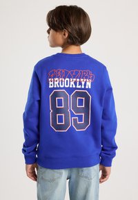 Bambino con capelli corti castani che indossa un felpa blu con la scritta "Brooklyn 89" sulla schiena, abbinato a jeans azzurri, rivolto di lato.