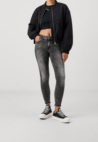 Persona que lleva una chaqueta bomber corta negra, un top corto negro, jeans ajustados gris descolorido y zapatillas negras de corte bajo con suelas blancas.