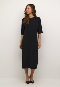 Abito nero a coste con maniche corte, lungo fino al ginocchio. Presenta un colletto rotondo e una silhouette aderente. Abbinato a scarpe nere con punta affusolata.