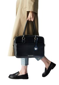 Borsa porta documenti nera in pelle con finiture dorate, forma rettangolare, tasca frontale e un portachiavi blu con un quadrante circolare. Persona vestita con jeans chiari e mocassini neri.