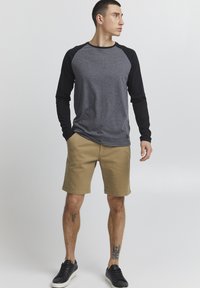 T-shirt à manches longues gris et noir avec un col rond, associé à un short beige ; le modèle porte des baskets noires.