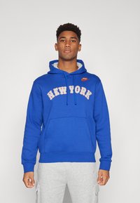 Zila Nike kapuce ar uzrakstu "NEW YORK" smilšu krāsā. Iespējas - priekšējā kabata un aukliņu kapuce. Valkāts ar pelēkiem biksēm.
