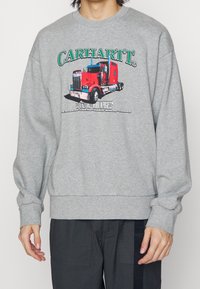 Šedá mikina s kulatým výstřihem, dlouhými rukávy a žebrovanými manžetami. Obsahuje barevný grafický potisk červeného trucku a text "CARHARTT".