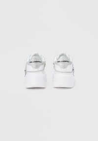 Λευκά αθλητικά παπούτσια με χοντρή σόλα, διακοσμημένα με ασημένιες λεπτομέρειες και γκλίτερ. Στο πίσω μέρος εμφανίζεται η μάρκα "KARL LAGERFELD".