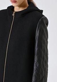 Cappotto nero con cappuccio, caratterizzato da un corpo in tessuto testurizzato e maniche in pelle trapuntata. Dettagli in oro per la cerniera e design minimalista.