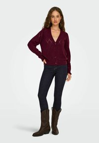 Cardigan en tricot bordeaux avec un col en V, un motif en losange et une fermeture boutonnée. Associé à un jean foncé et des bottes de cowboy marron.