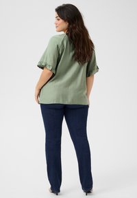 Top corto a maniche corte verde oliva con orli arricciati, abbinato a jeans dritti blu scuro. Tessuto morbido, design casual.