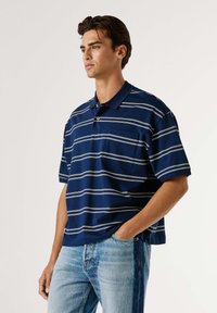 Mornarsko modra polo majica z belimi vodoravnimi črtami, kratkimi rokavi in ovratnikom. Ima žep na prsih in sproščen kroj. Oblečena je z svetlo modrimi kavbojkami.