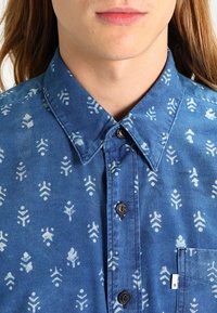 Blauwe denim shirt met witte gedrukt patronen, een gespreide kraag, knoopsluiting en een borstzak. Gelaagd stof met een zachte afwerking.