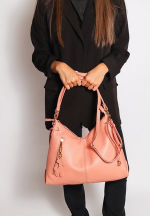 Femme en tenue noire tenant un grand sac à main rose texturé avec une poche zippée et un porte-clés en forme d'ours en peluche.