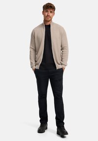 Lichtbeige zip-up jack met textielstructuur, gedragen over een zwart shirt, gecombineerd met donkere broeken en zwarte schoenen.