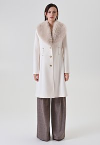 Cappotto di lana bianco con un ampio colletto di pelliccia beige, bottoni dorati e coulisse regolabili. Abbinato a pantaloni larghi a quadri con motivo.