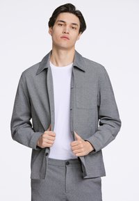 Graue, strukturierte Jacke mit spitzem Kragen und Brusttasche, getragen über einem weißen T-Shirt; verfügt über eine Knopfverschluss und eine lässige Passform.