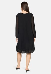 Czarna plisowana sukienka z długimi, przezroczystymi rękawami, okrągłym dekoltem i długością do połowy łydek. Miękka, lekko płynąca tekstura podkreśla ruch.