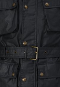 Musta vedenpitävä takki vyöllä, messinkisillä napeilla, kahdella rintataskulla ja teksturoidulla kankaalla, jossa on jäsennelty muotoilu ja selkeät linjat.