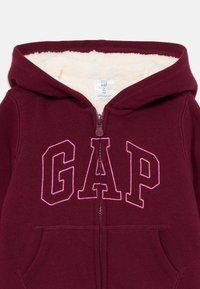 Bordový detský mikinový sveter s bielym fleecovým podšívaním, predným zipsom a ružovo obkresleným vyšitým logom "GAP" na hrudníku.