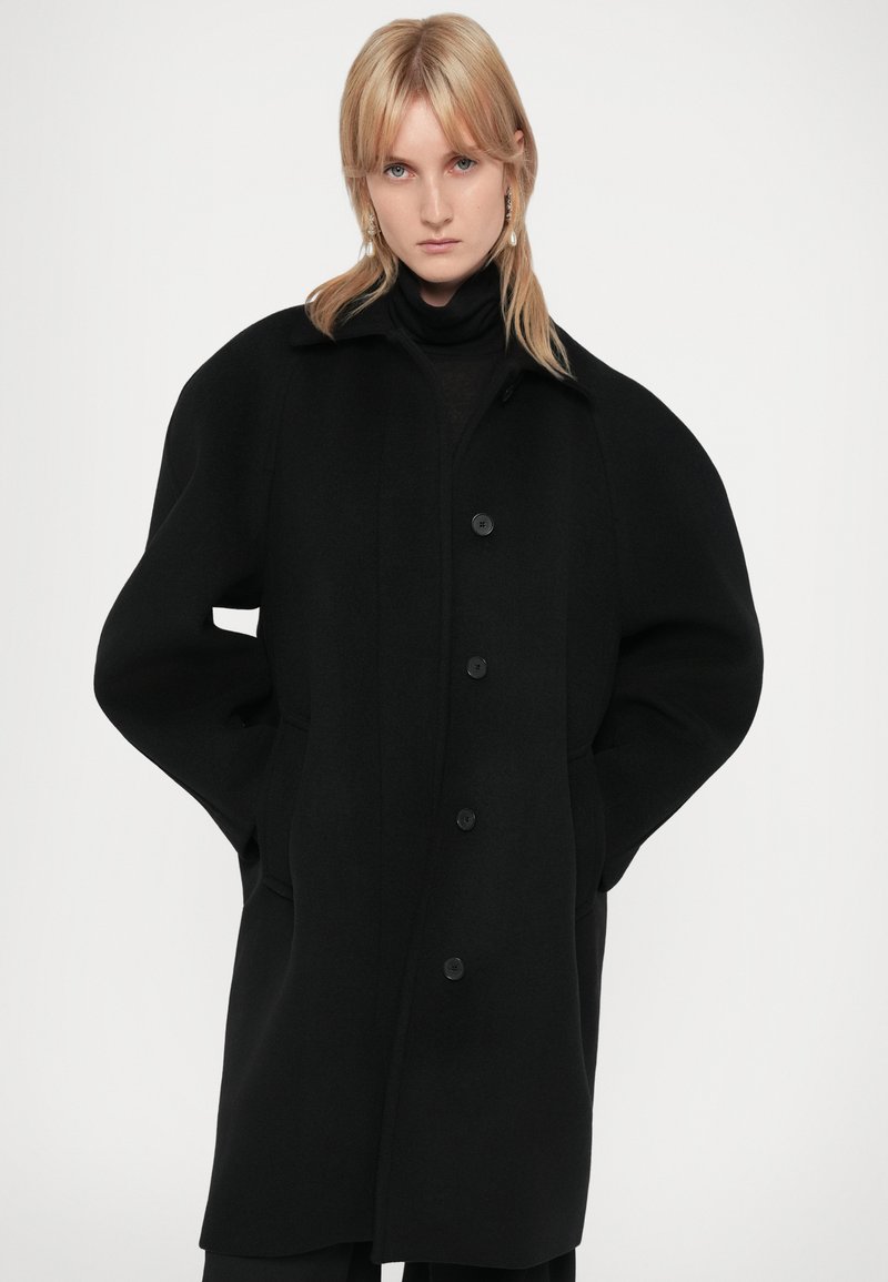 Svart ullkappa med rak design, överdimensionerade axlar, stora knappar och sidofickor. Textilerad tyg med en strukturerad siluett.