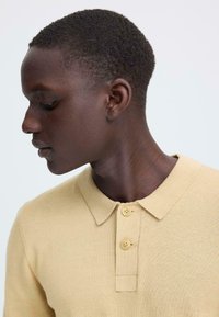 Polo en maille beige clair avec une texture douce, doté d'un col et de deux boutons sur la patte de boutonnage, porté par une personne de profil.