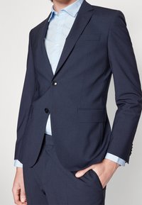 Midnattsblå kavaj med smal passform, enkel knappstängning, slitsade slag och två framfickor, kombinerad med en ljusblå skjorta med krage.