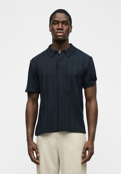 Άνδρας που φοράει σκούρο πουκάμισο πόλο με σχέδια και κοντά μανίκια, καθώς και ανοιχτό μπεζ παντελόνι, στέκεται μπροστά σε απλό λευκό φόντο.