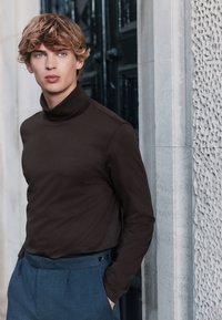 Brun polotröja i mjukt tyg, kombinerad med skräddarsydda blå byxor med knappränder. Modellen står mot en strukturerad vägg.