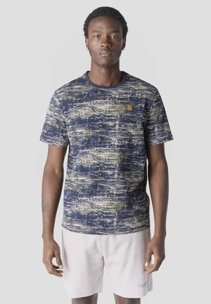 Mladý muž oblečený v modro-bežovej vzorovanej košeli s krátkym rukávom a bielych šortkách, stojaci pred jednoduchým svetlosivým pozadím.