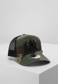 Camouflage-Kappe mit einem schwarzen bestickten Logo, Mesh-Rücken, gebogenem Schild und verstellbarem Riemen. Der Stoff ist eine Mischung aus grünen und braunen Farbtönen.