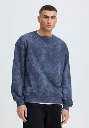 Homme portant un sweat-shirt à col rond bleu à motifs et un pantalon noir, debout devant un fond clair uni.