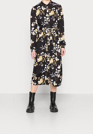 Zwarte bloemenjurk met lange mouwen, knoopsluiting aan de voorkant en tailleband. Heeft gele en witte bloemenpatronen. Gecombineerd met zwarte enkellaarsjes.