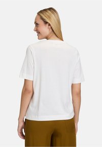 T-shirt en coton blanc à manches courtes, légèrement ample, avec un petit détail circulaire au dos du cou. Associé à un pantalon marron.