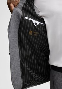Grau melierter Wollblazer mit schwarzem gestreiftem Innenfutter, versehen mit einem Etikett, das "Manteco Wool" liest, und einem weißen Einstecktuch.