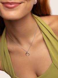 Femme portant un haut vert à col halter et un collier en forme de croissant de lune argenté, souriant doucement avec des lèvres et un cou visibles.