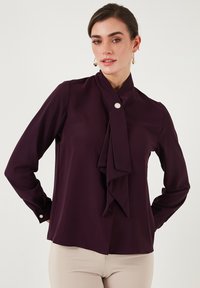 Blusa viola con una parte anteriore arricciata e chiusura con bottoni. Maniche lunghe con polsini abbottonati, realizzata in un tessuto leggero e liscio.