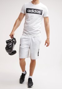 Camiseta gris de Adidas con logo negro, pantalones cortos blancos con detalles negros, sosteniendo guantes de boxeo negros, vistiendo zapatillas deportivas negras.