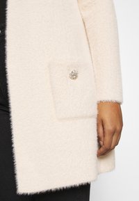 Krémový kabát s texturou a jemným povrchem, vybavený našitou kapsou s ozdobným knoflíkem z broušeného kamene. Černá látka pod ní.