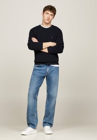 Maglione girocollo navy con tessuto testurizzato sovrapposto a una t-shirt bianca, abbinato a jeans dritti azzurri chiari e sneakers bianche.