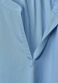 Světle modrá látka s hladkou strukturou. Výstřih do V s malým trojúhelníkovým výřezem ve středu. Měkký, splývavý design.