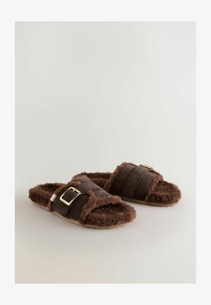 Pruun karvane suss, millel on kaks tekstuuritud rihma, üks kuldse metallist pandlaga ja pehme tallaosa mugavuse tagamiseks.