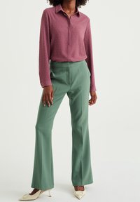 Maroonin teksturoitu paita, jossa on kaulus, yhdistetty vihreisiin räätälöityihin levennettyihin housuihin. Mallilla on päällä kermaiset korot, joissa on yksityiskohtia edessä.