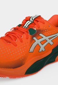 Φωτεινό πορτοκαλί αθλητικό παπούτσι Asics με λευκό και μαύρο λογότυπο, κόκκινα κορδόνια και μαύρες λεπτομέρειες στη σόλα και την επάνω πλευρά.