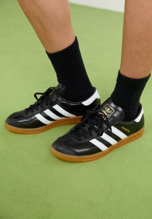 Pieds portant des baskets Adidas Hamburg noires avec des bandes blanches et des semelles en gomme, assorties à des chaussettes noires côtelées sur un sol vert.