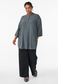 Lagani, košulja na dugmad u tamnosivoj boji s mandarin kragnom i rukavima do tri četvrtine, u kombinaciji s širokim crnim hlačama i cipelama na životinjski uzorak.