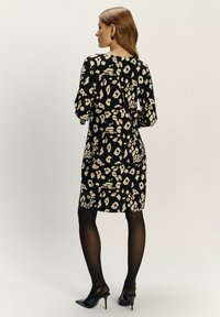 Zwarte jurk met beige dierenprint, lange mouwen, knie-lengte en een rechte pasvorm. Draagt met zwart doorschijnend panty en hoge hakken.