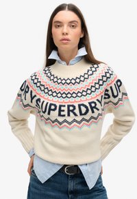 Suéter de punto crema con patrones geométricos en azul marino, coral y azul claro. Presenta la palabra "SUPERDRY" de manera destacada en el frente.