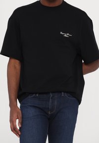 Černé tričko s krátkými rukávy, volný střih, kulatý výstřih a vyšívané logo bílé barvy na levé hrudi. Džínové kalhoty viditelné dole.