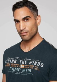 Mörkblå v-ringad t-shirt i mjukt tyg, med vita och orange textgrafik, inklusive en logotyp och designelement på framsidan.